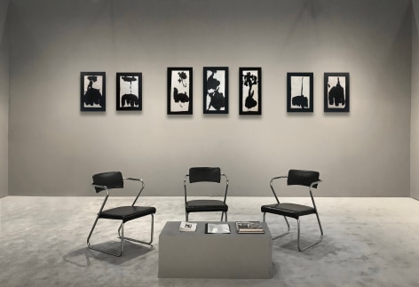 Seven works on paper by Richard Stankiewicz in expressive strokes using black ink on white paper hanging on a grey wall.  Three chairs and a table in the foreground.