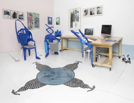 A photograph of the back office of the gallery that shows the desks and unique chairs created by the artists out of blue duct tape. On the walls are numerous mixed media works in natural frames. On the ground is the site-specific installation with black and white triangles.