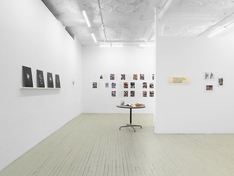 A photograph of the gallery with 4 black prints on the left wall on a shelf, leaning; a table with items on it in the middle-ground; a group of 4 items unframed on the temporary wall at right; and an excerpt of an artwork containing magazine spreads on the back wall.