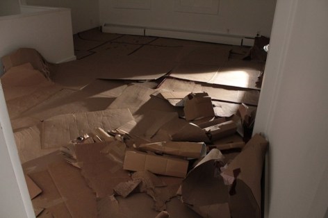 A photograph of the gallery floor. There is nothing on the wall, and panels of cardboard spread throughout the floor. They are used, and there is a pile at right of cylinders/broken boxes