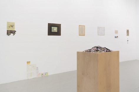 An installation view that includes small magazine clippings piled on a wooden pedestal in the foreground, and 6 mixed-media works installed on the wall.