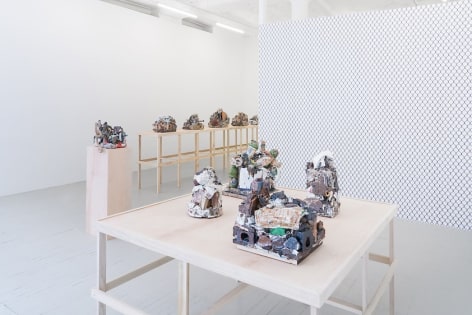 A photograph providing the view of the gallery from the front window. The square raw table with 4 ceramic sculptures is in the foreground, a single pedestal with a ceramic sculpture is in the midground, and in the background we see a long raw platform with 4 visible sculptures upon it. We also see the chainlink wallpaper on the temporary wall jutting from the right of the photo.