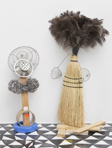 A photograph of two mixed media sculptures: on the left is a work made of tape rolls, steel wool, and 2 colanders; at right is a sculpture made of the head of a broom and a feather duster at the top with two small strainers jutting from either side.