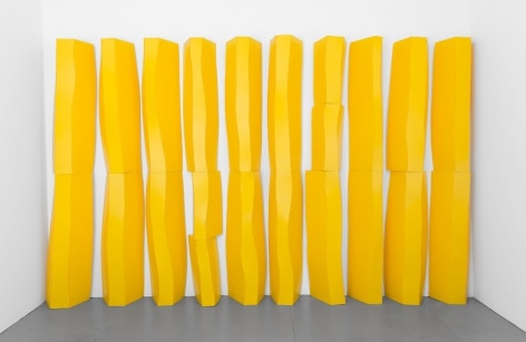 A photograph of an installation of tall geode-like shapes stacked upon one another. There are 10 columns of these 3D, yellow shapes. They appear like cartoonish rocks or glaciers.