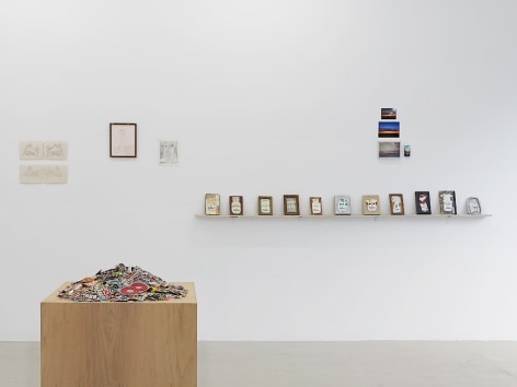 An installation view with a pedestal filled with magazine clippings in the foreground at left. On the wall in the distance is a sculpture made up of old cigarette cartons on a shelf, and 2 other drawings on paper without frames.