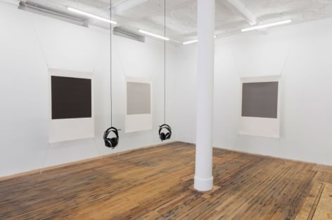 A photograph of the back corner of the gallery, where 3 screenprint works by Bergvall are stretched at all 4 of their corners. In the center of the room are 2 hanging sets of headphones.