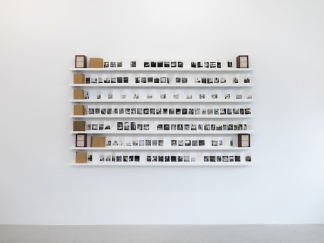 An installation view of &quot;Sent a Letter&quot; where each of the 7 accordion photo-books are unfolded and visible on a white shelf hung on the wall. You see 4 of the brown leather cases that surround the linen box, meant to hold all 7 books.