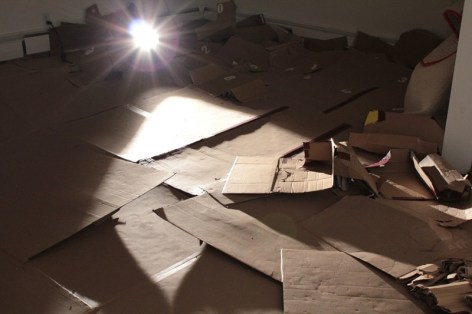A photograph of the gallery floor. There is nothing on the wall, and panels of cardboard spread throughout the floor. They are used. A projector is at top-left of image
