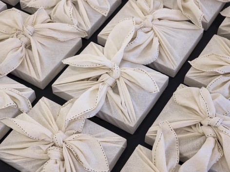 A photograph of the exterior of Dayanita Singh's Pothi Boxes, which are wrapped in linen napkins with 2 pothi knots visible. The boxes are laying on a black table, and the image is a close-up of 9 boxes.