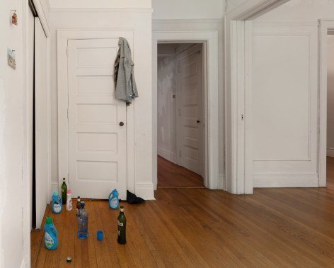 A photograph of the left side of the installation with detergent and liquor bottles on the ground, a shirt over a door in the background