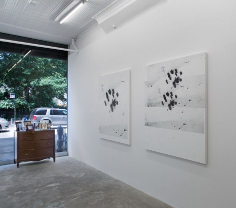 A photograph of the front of the gallery: at right are two white paintings with black cat paws; near the window we see a wooden dresser with illegible photographs upon it.
