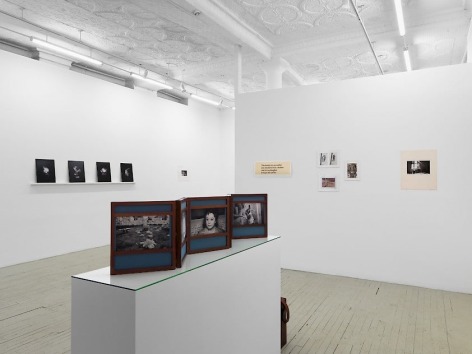 A photograph of the gallery. We are standing behind Dayanita Singh's unfurled book-object upon a pedestal. In the background are 5 works on the temporary wall at right, and a series of 4 black prints by Pradeep Dalal on the left side. In the distance at left is a photograph by Guibert that is illegible.