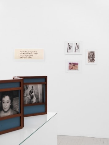 A photograph that details 4 works on the temporary wall. The work at left has 4 lines of text. To the right are three color photographs hung in a loose triangle.