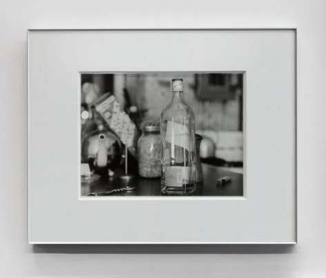 A black and white photograph of a bottle on a table amid other kitchen accessories.