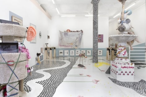 A photograph of the interior of the gallery. There is a site-specific installation with black triangles on the ground and flowers painted to be coming out of it. There are several mixed media sculptures installed on the ground. On the walls at more mixed-media works with the contents illegible. The pole is painted black and white with triangles.