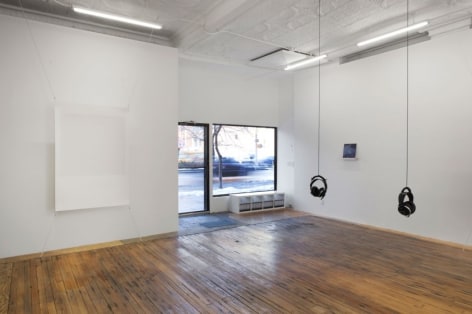 A photograph of the interior of the gallery, with a view of the windows in the background. There are 2 sets of headphones hanging from the ceiling, and Tom Martin's piece at right in the background. At left is a screenprint stretched at 4 corners, off the wall.