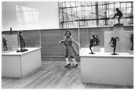 A black-and-white photograph of a young Black girl dancing in a museum, where bronze sculptures are contorted within vitrines
