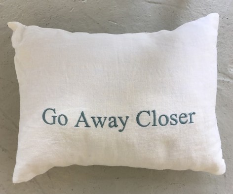 A white pillow with &quot;Go Away Closer&quot; embroidered in grey-blue.