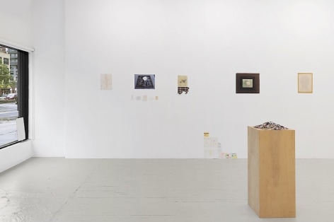 An installation view that includes small magazine clippings piled on a wooden pedestal at right, and 4 mixed-media works installed on the wall.