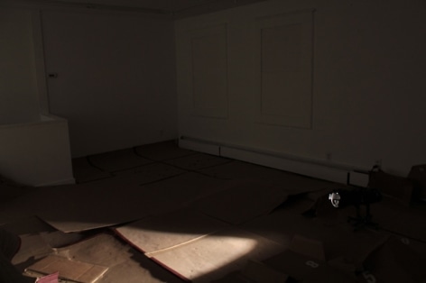 A photograph of the gallery floor. There is nothing on the wall, and panels of cardboard spread throughout the floor. They are used, the room is mostly dark