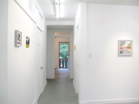 A photograph of 3 artworks: one on the wall facing us, and 2 on the wall of a hallway. At the end is a doorway to the outside