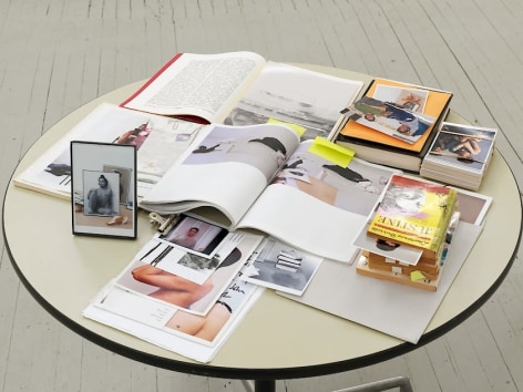 A round cream-colored table covered with the items described in the artwork's caption. They are layered upon one another, some invisible beneath piles of paper.