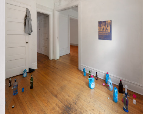 A photograph of an installation in an entry foyer, with a shirt over a door, open detergent and liquor bottles, and a work on the wall at right