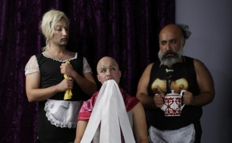 A freeze-frame of a film work. The image depicts the three artists. At left, the artist is dressed as a traditional french maid. The figure in the middle holds toilet paper in his mouth. The figure at right is wearing a french maid bib with a t-shirt.