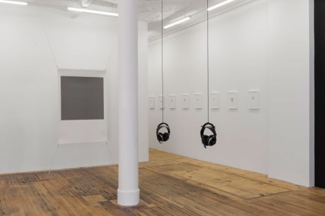 A photograph of the view of the back quadrant of the gallery. There is one screenprint with a gray center, 2 sets of headphones hanging in the center of the room, and a series of smaller works on the wall framed in white. Those works run in a row toward the back office.