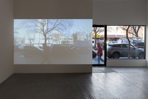 A photograph from the inside of the gallery. On a white wall is a projection of the sidewalk. To the right of the projection is the front window of the gallery, which depicts the sidewalk and street traffic on Delancey Street.