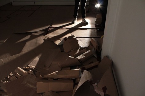 A photograph of the gallery floor. There is nothing on the wall, and panels of cardboard spread throughout the floor. They are used.