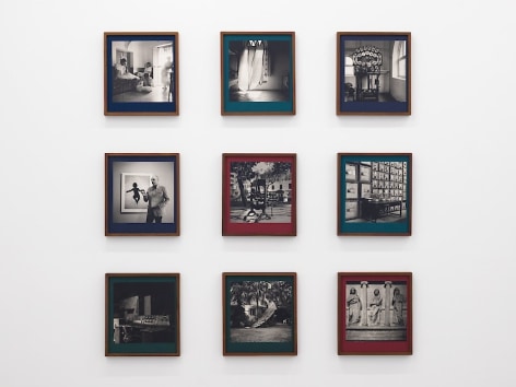 A 3 x 3 grid of 9 books, each contained in a teak wood frame. They have varied colors of linen (crimson, teal, green, navy) and each book has a different black and white photograph on the cover. They are hung on a white wall.