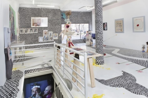 A photograph of the gallery from the back corner looking toward the door. A view of the basement staircase, showing a video at the bottom, is at left. The rest of the room is filled with flat works installed on the wall, mixed media sculptures installed on the ground, and a site-specific installation of black and white triangles on the ground and walls.