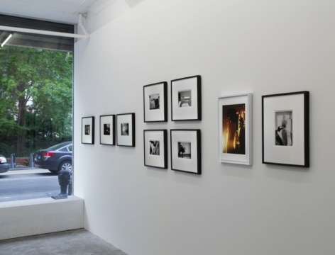 A photograph of 9 images on a white wall. At left is the window of the gallery looking outside