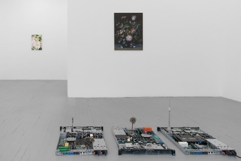 A frontal view of the gallery, with the circuit board sculpture in the foreground. Just behind it we see a still life painting on black ground, and in the distance is an abstract, multi-colored painting.