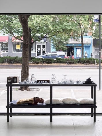 An installation view of a shelf containing various glass items, 3 pillows, and tote bags