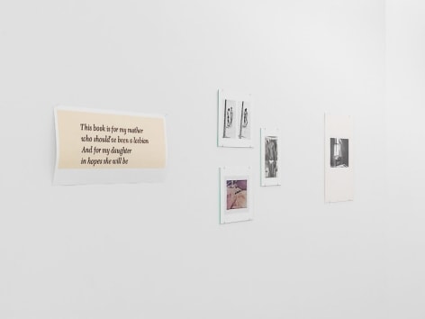 A photograph of the artworks on the temporary wall: there is a single work with text, 3 works hung in a loose triangle to the right, then a single photograph by Guibert of a man hunched over a desk with light streaming through a window above his head.