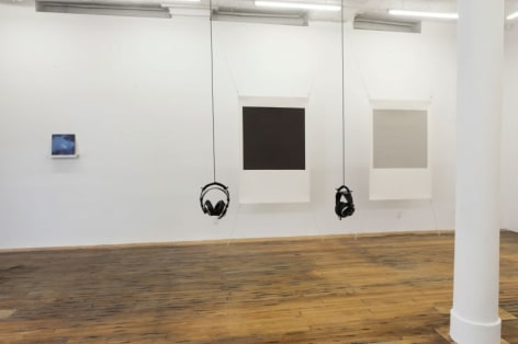 A photograph of the interior of the gallery. There are 2 sets of headphones, Tom Martin's piece at left on the wall in the background, and 2 screenprints by Bergvall stretched at the four corners on the same wall.
