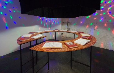 An installation of 15 books on a circular wooden table, some open and some closed. There are red, green, and red LED lights on the walls.