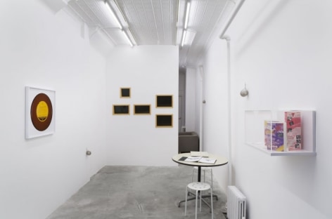 A photograph of the gallery with a sparse install: a table on the right, a vitrine on the right wall, 5 black rectangles on the back wall, and a framed record at left.