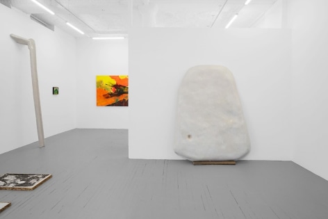 A photograph of the gallery where we see the temporary wall with a large white sculpture upon it. There is a partial view of a colorful painting on the back wall and a smaller painting at left of that. There is a large sculpture leaning against the wall at left, and a print installed on the ground near the left-corner of the photograph.