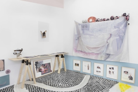A photograph of the gallery's site-specific installation. At right is a work on fabric with small sculptures above it. At left is a table made of plywood and 2 sawhorses that hold more flat works. There are also works beneath the table at left and right, framed in natural wood. Contents are illegible.