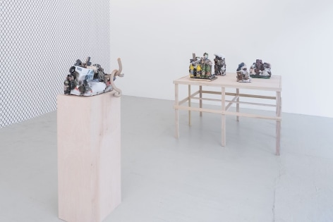 A photograph of a single raw wood pedestal with a ceramic sculpture upon it. In the background we see a raw wood platform with 4 ceramic sculptures upon it. At left, we see the chainlink fence wallpaper on the gallery's temporary wall.