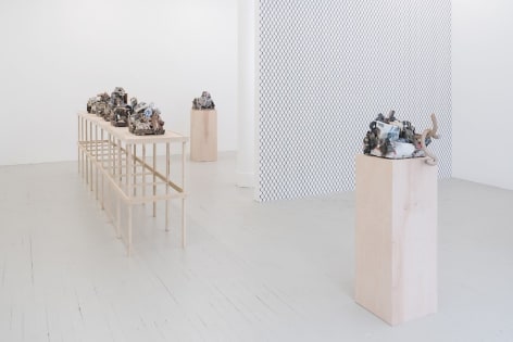 A photograph of the long raw wood platform holding 5 ceramic sculptures, and 2 sculptures on raw wood pedestals (1 each respectively), one in the foreground, one in the background. The chainlink fence wallpaper is visible in the middle ground.