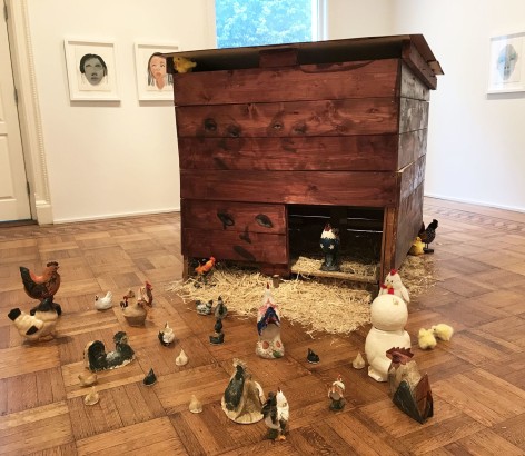 Installation work by February James consisting of a wooden chicken coop with faces painted on each side and tens of found ceramic chickens on the floor.