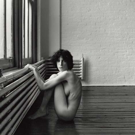 Nude Patti Smith with dark tousled hair sitting curled up in profile while holding on to a radiator by a window in a room with dark hardwood floors and white brick walls. Her head is turned to look directly at the viewer.