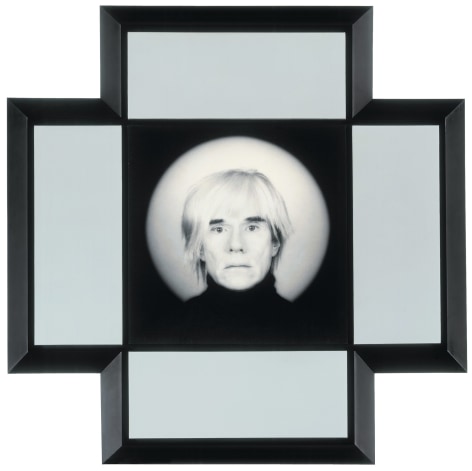 A portrait of the artist Andy Warhol from the neck up in a black turtleneck looking at the viewer. The black frame with white mat around the image forms a cross shape.