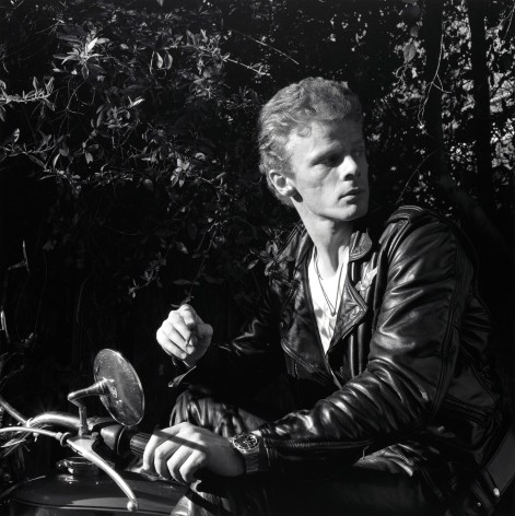 Man wearing a leather jacket and smoking sitting on a motorcycle outdoors in front of foliage.