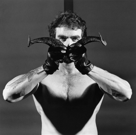 Shirtless man wearing leather gloves holding two horns in front of his face.