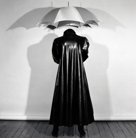 Back view of a woman wearing a long leather jacket holding an umbrella above her head and casting shadows on the white wall before her.